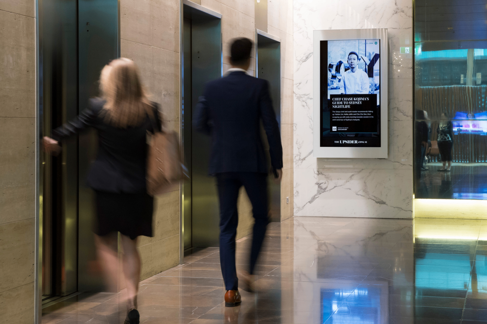 Digital screen in a marble office tower lobby beside elevator banks — the kind of in-building inventory ABM campaigns place to reach decision-makers at their actual workplace