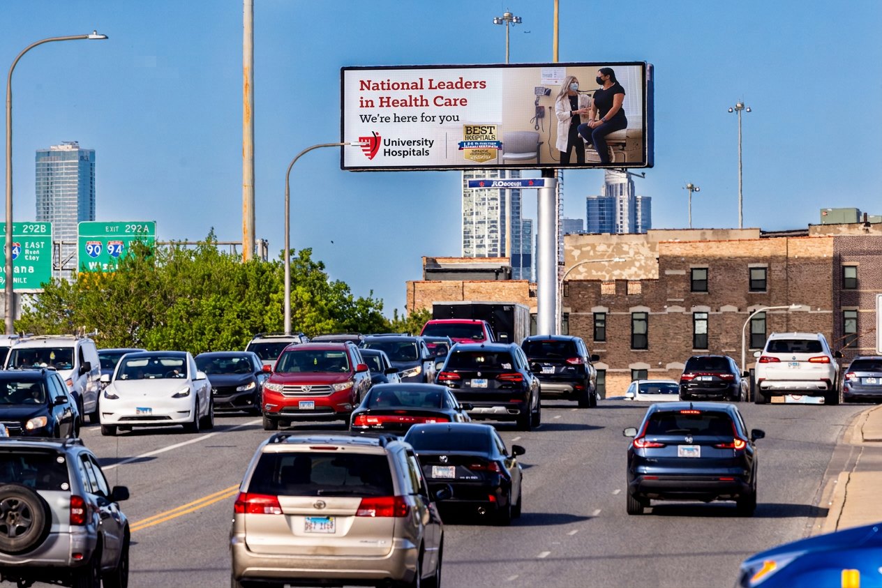 A digital billboard mockup over a highway showing a University Hospitals brand ad reading National Leaders in Health Care, with a city skyline behind it.
